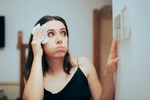 Woman sweating as she looks at thermostat because AC is blowing warm air.